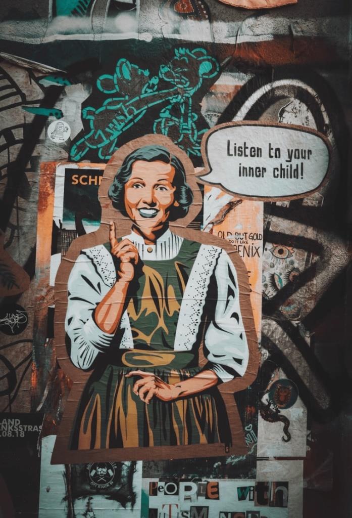 The image shows wall art. A woman is dressed in 1950’s style. A speech bubble next to her says, ” Listen to your inner child” There are cartoons and graffiti on the wall too